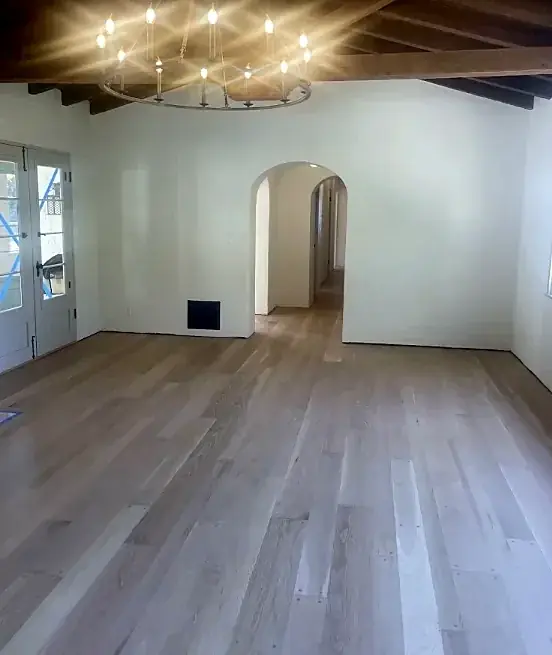 A white room with dark beams, an iron chandelier and an arched doorway, and a pale natural wood floor