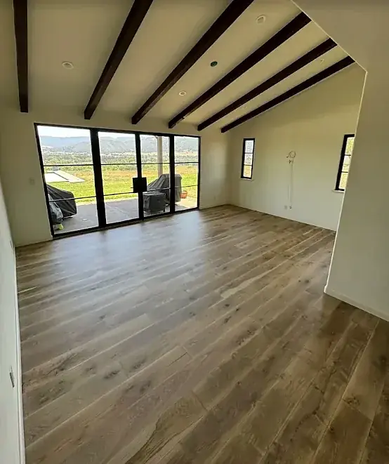 A beamed room with a gray-brown wide plank floor and black-framed glass doors to a patio and hills