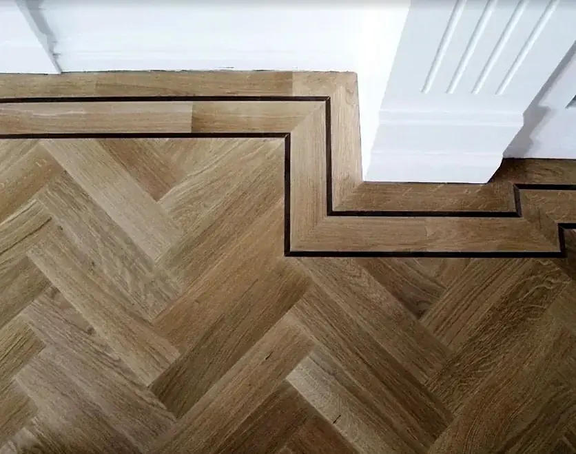 A herringbone floor with a dark inlaid line that steps around the base of a wall