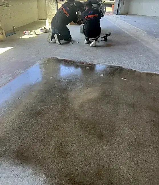 Two crew members in La Cumbre shirts kneel at the edge of a wet coating spread on a concrete floor