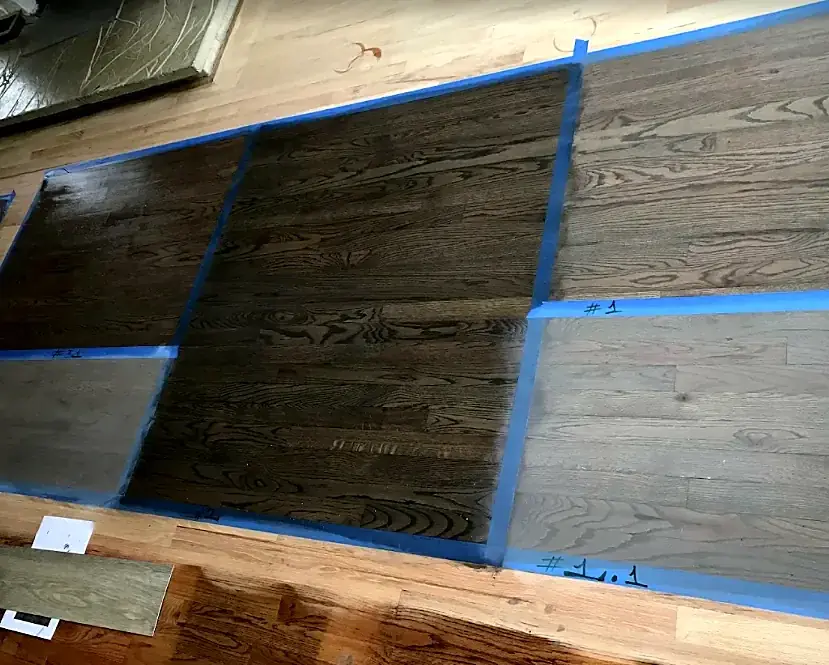 Dark and gray stain samples in taped boxes on a sanded wood floor