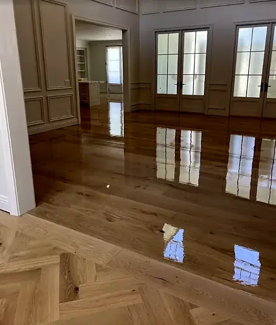 A paneled room with wide plank flooring reflecting three French doors, and a herringbone floor in the doorway in front