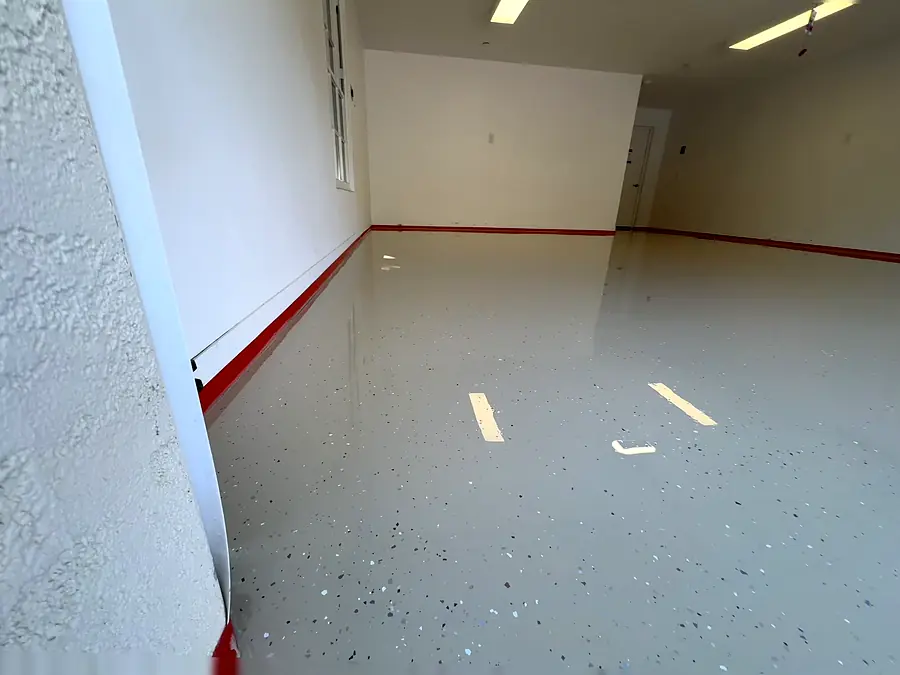 A wet epoxy coat in a garage with dark flakes scattered across it, red tape along the wall and blue paper at the edge
