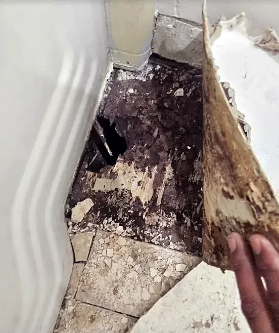 A hand peels back old flooring in a corner to show dark, crumbling material underneath