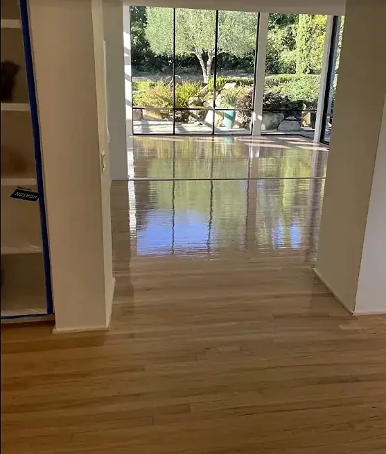 A refinished oak floor with a high gloss runs through an opening to a wall of windows on a garden