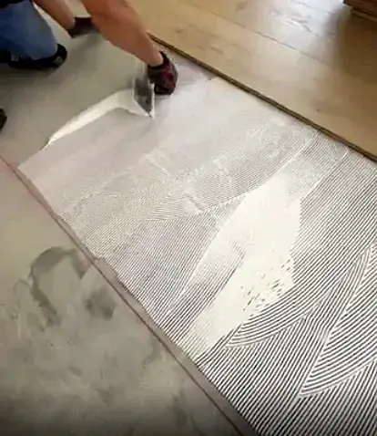 A gloved hand combs white adhesive into ridges with a notched trowel, next to the last row of planks