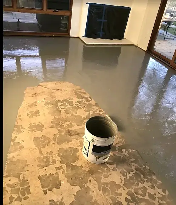 Wet gray leveling compound spreads across a room around a patch of old slab, with an empty bucket in the middle