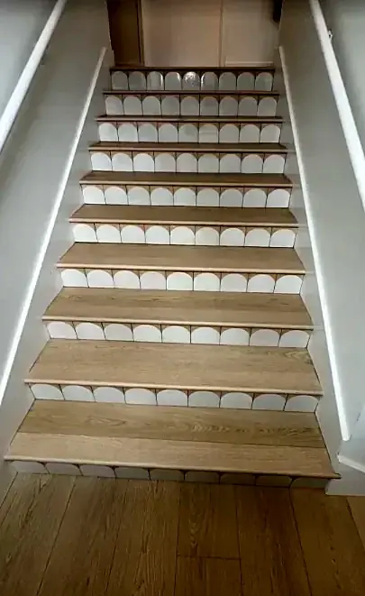 A straight stair seen from the bottom, with light oak treads and scalloped white tile risers