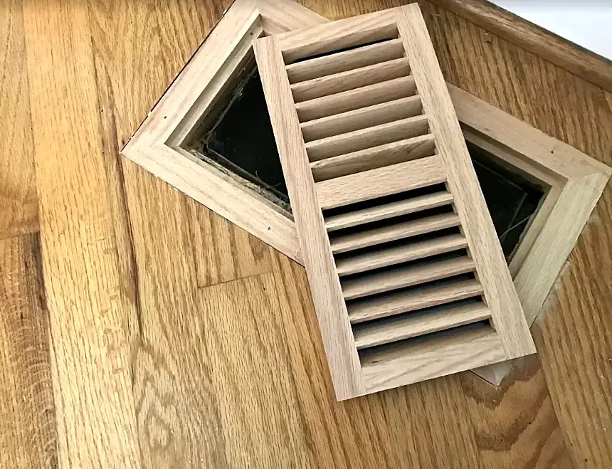 An unfinished wood vent frame and louvered insert resting on an oak floor over the vent opening