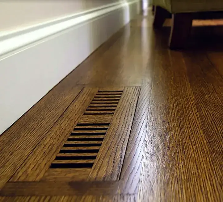 A wood floor vent set flush into a stained oak floor, its slots running with the boards